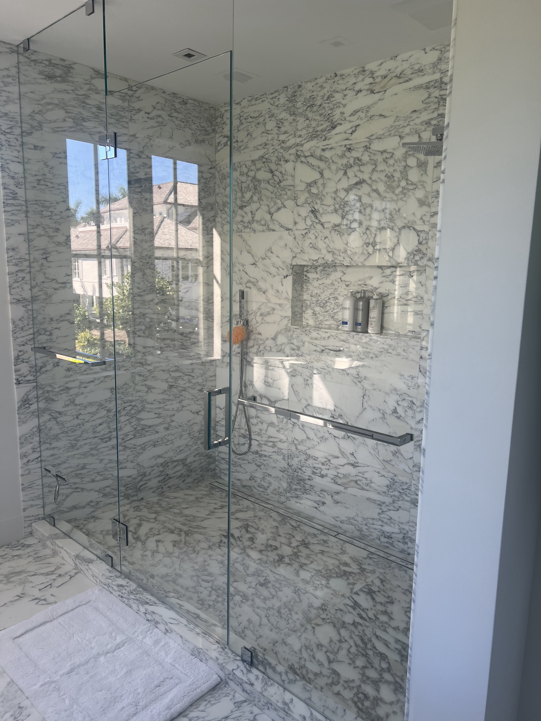 Sunlit frameless glass shower with white marble walls and chrome hardware